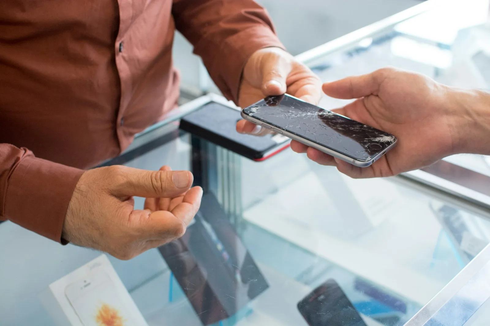 Customer handing a smartphone with a cracked screen to a repair shop technician over the counter, illustrating the moment of intake