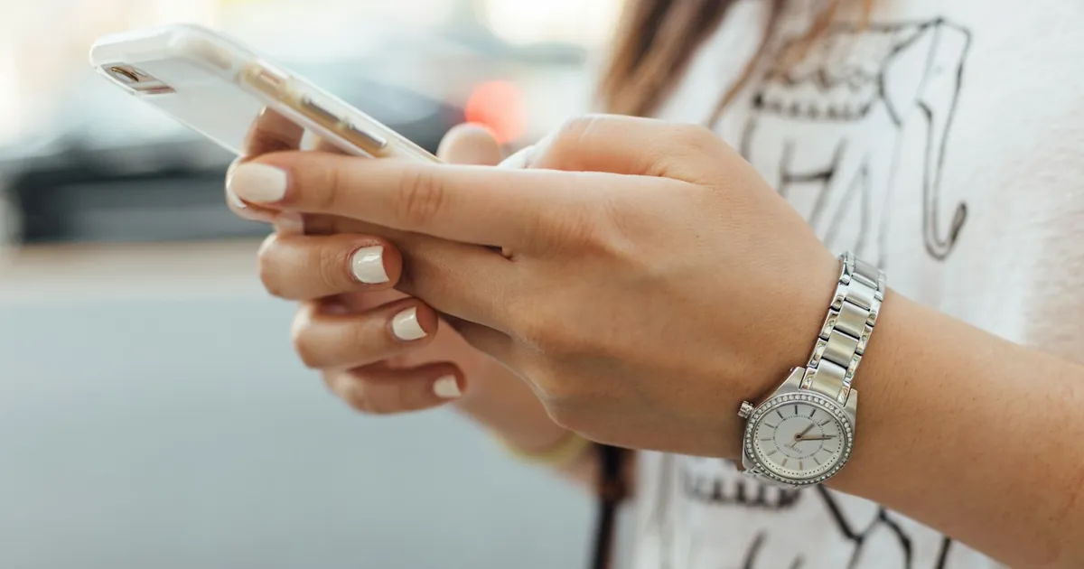 A woman with a wristwatch holds a smartphone in both hands and types a text message outdoors.